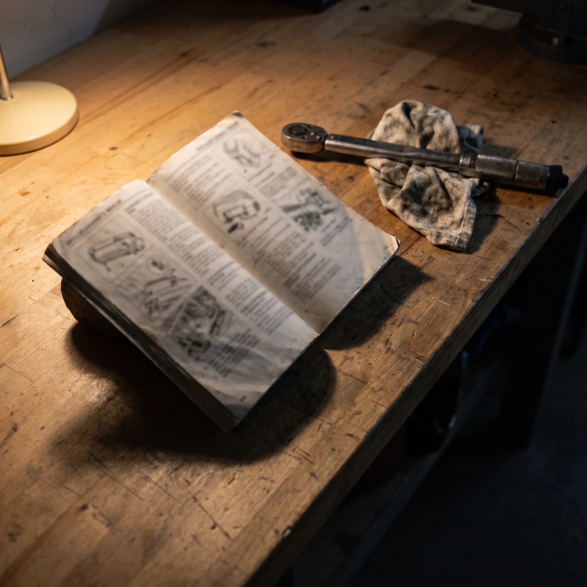 Open vehicle owner's manual on a worn workbench next to a wrench and a rag under a single work lamp